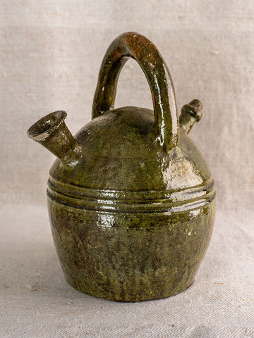 Antique French green glazed earthenware gargoulette jug with round body and two spouts on neutral fabric background.
