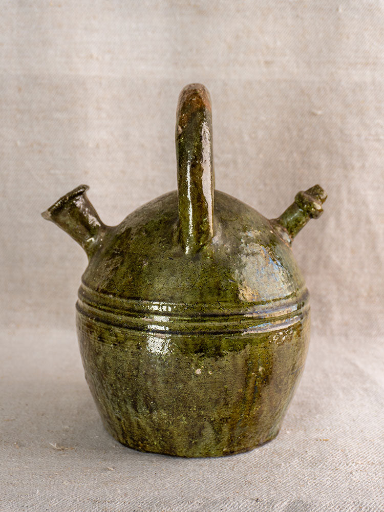 Antique French green-glazed earthenware gargoulette, c.1850, with upright handle and two spouts on neutral fabric background.