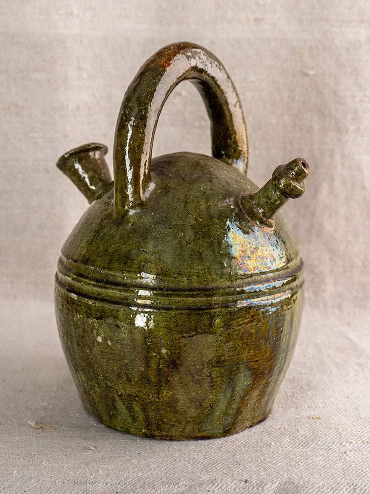 Antique French green glazed earthenware gargoulette jug with dual spouts and handle on textured fabric.