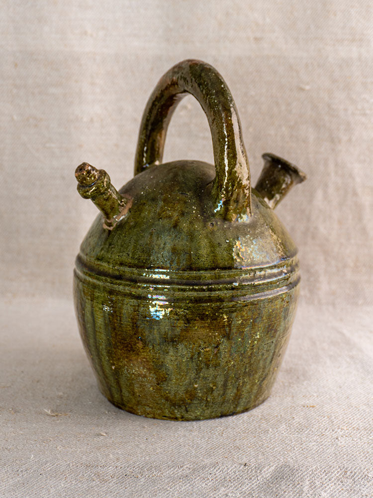 Antique French green-glazed earthenware gargoulette with curved handle and twin spouts on beige textured cloth.