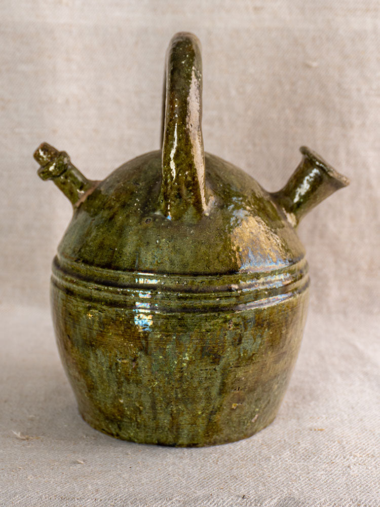 Antique French green glazed earthenware gargoulette with arched handle and twin spouts on beige textured fabric.