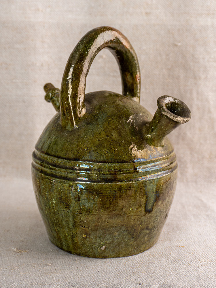 Antique French green-glazed earthenware gargoulette with round body, arched handle, and angled spout on neutral fabric.