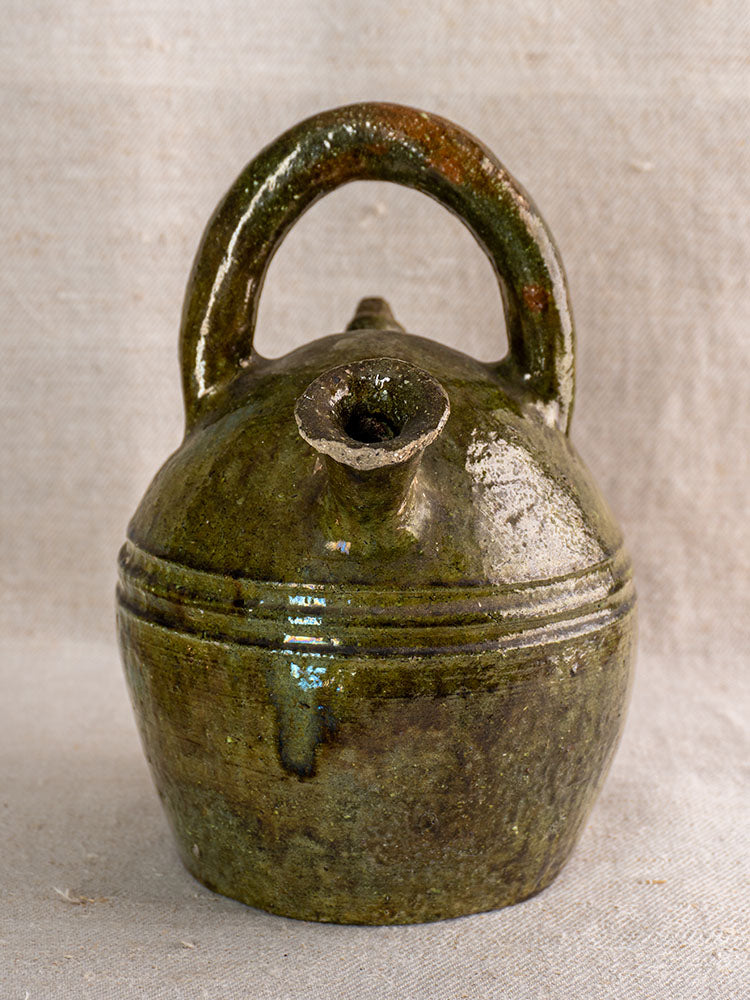 Antique French green glazed earthenware gargoulette jug with rounded body, curved handle, and spout on neutral fabric background.
