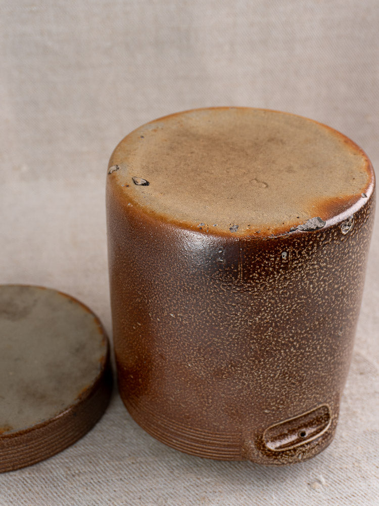 Antique French stoneware grease pot and matching salt glaze lid on textured beige fabric.