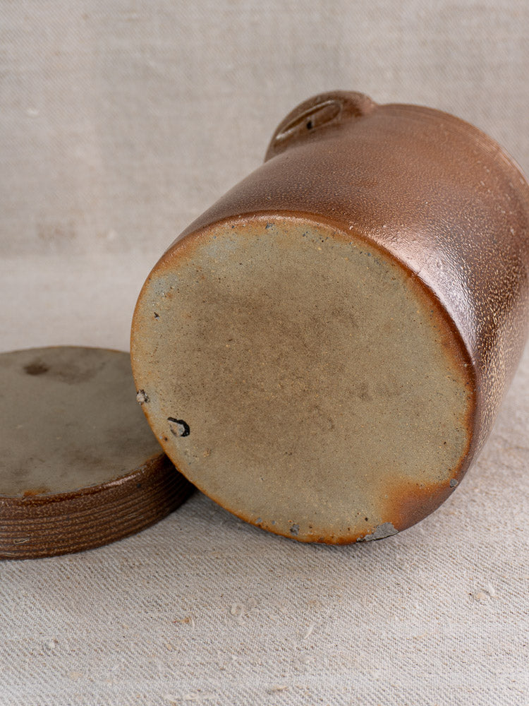 Antique French stoneware grease pot with salt glaze and matching lid on beige textured fabric.