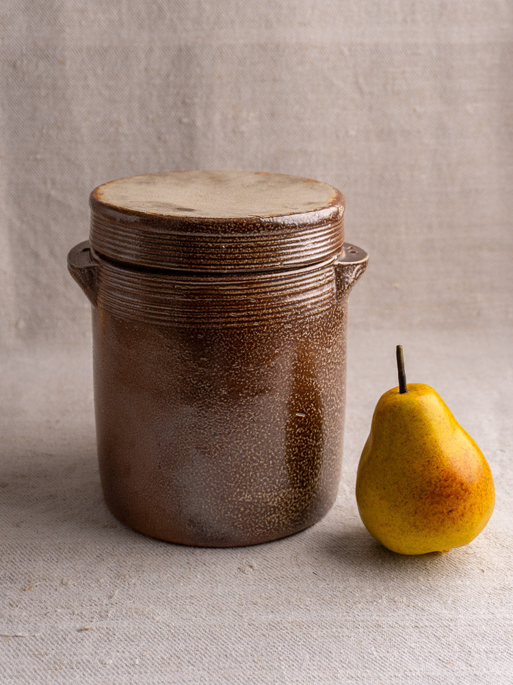 Antique French stoneware grease pot with matching salt-glaze lid beside a yellow pear on beige fabric.
