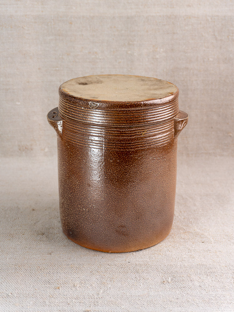 Antique French stoneware grease pot with brown salt glaze, light lid, and side handles on fabric.