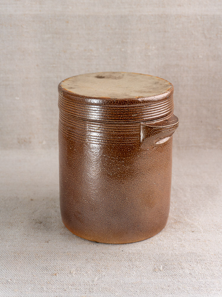 Antique French stoneware grease pot with salt glaze, matching lid, and side handle on fabric background.