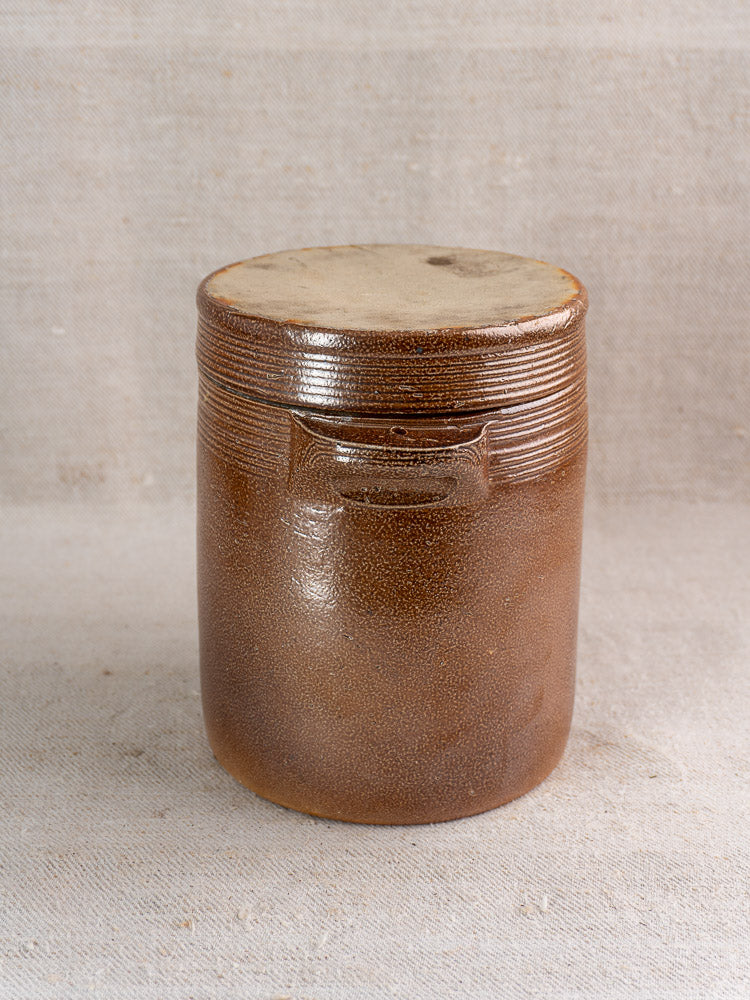 Antique French stoneware grease pot with salt glaze and lid on textured neutral fabric.