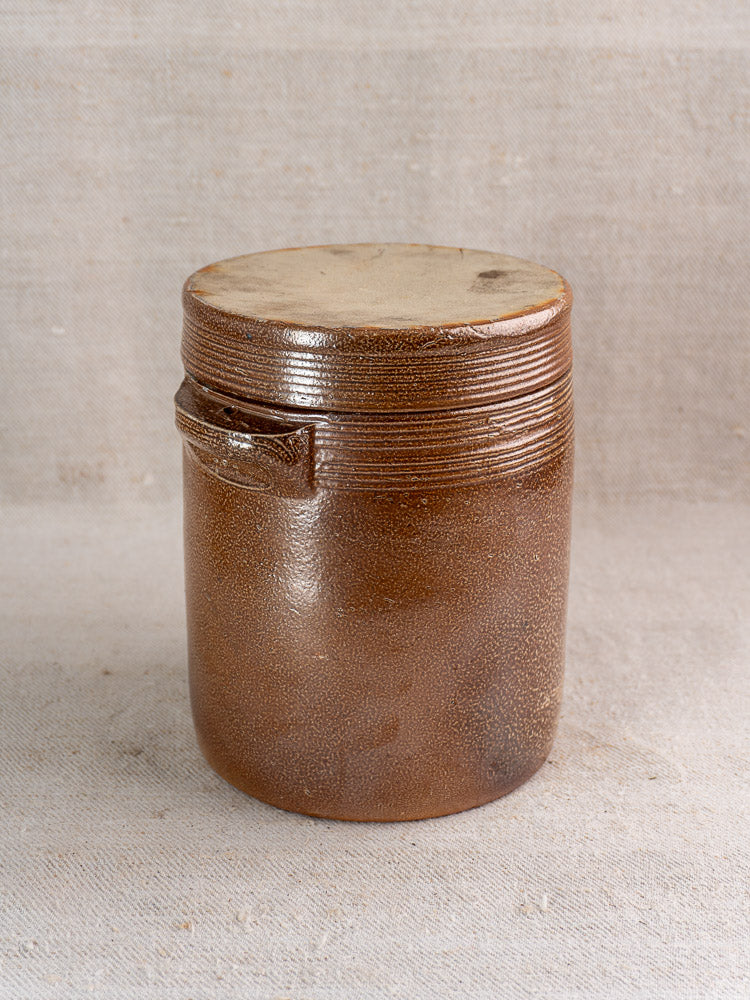 Antique French stoneware grease pot with matching lid and side handle on beige fabric background.