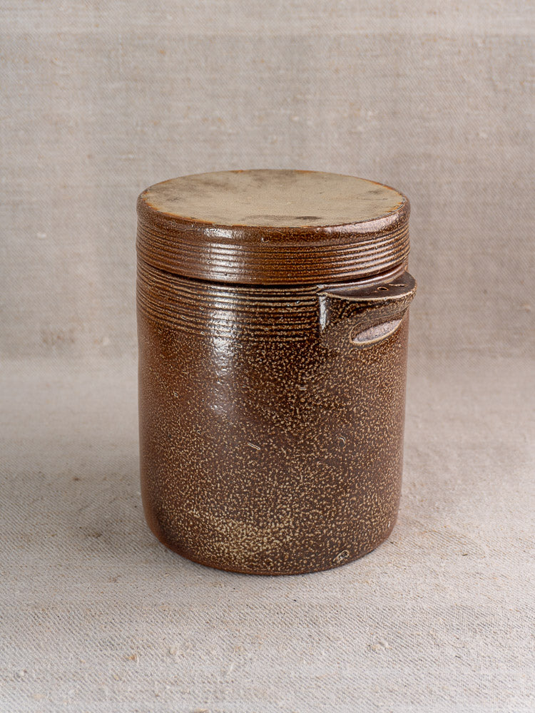 Antique French stoneware grease pot with salt glaze, brown textured body, side handle, and lid on neutral fabric.