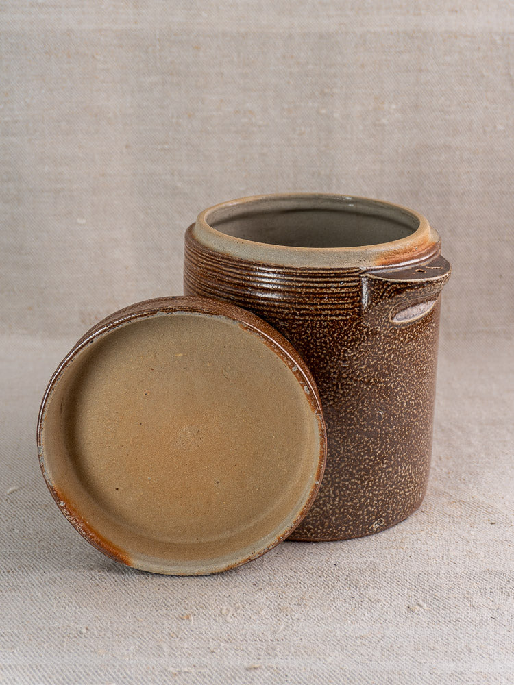 Antique French stoneware grease pot with brown salt glaze, textured surface, and matching lid on beige fabric.