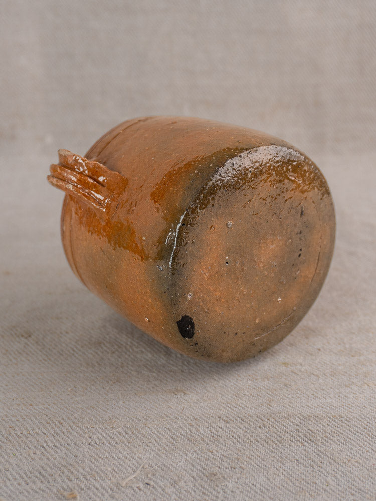 Antique French Vallauris pot with glossy brown glaze and side handle on beige textured fabric.