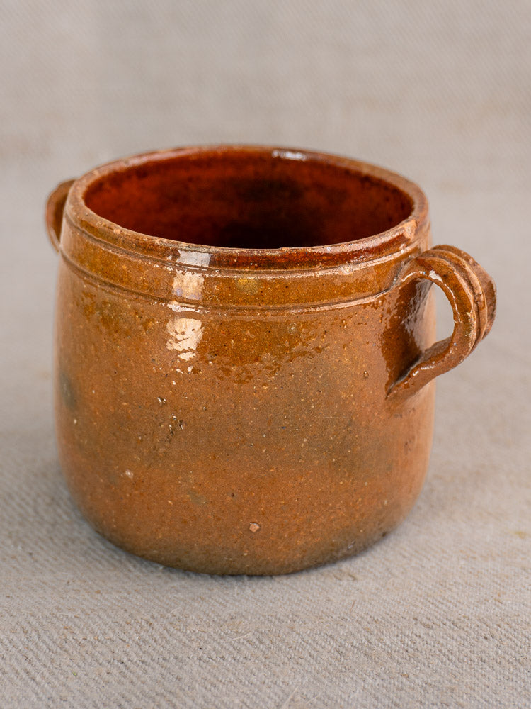 Small round brown glazed ceramic pot with two handles, from French Vallauris set, on gray fabric.