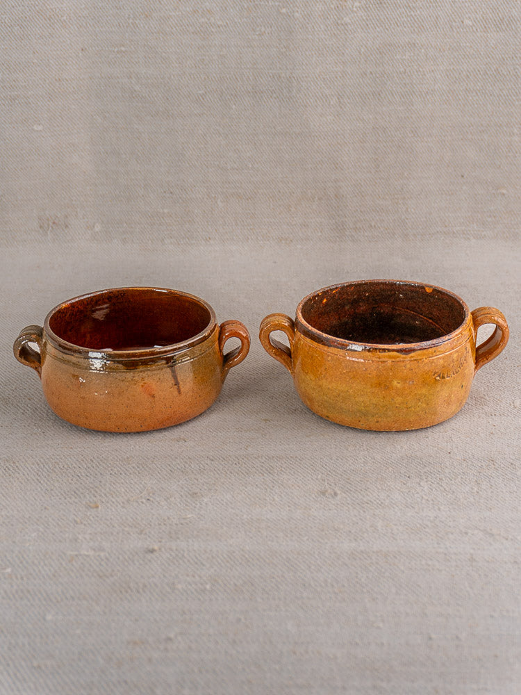 Set of three antique French Vallauris terracotta pots with side handles, on beige fabric surface.