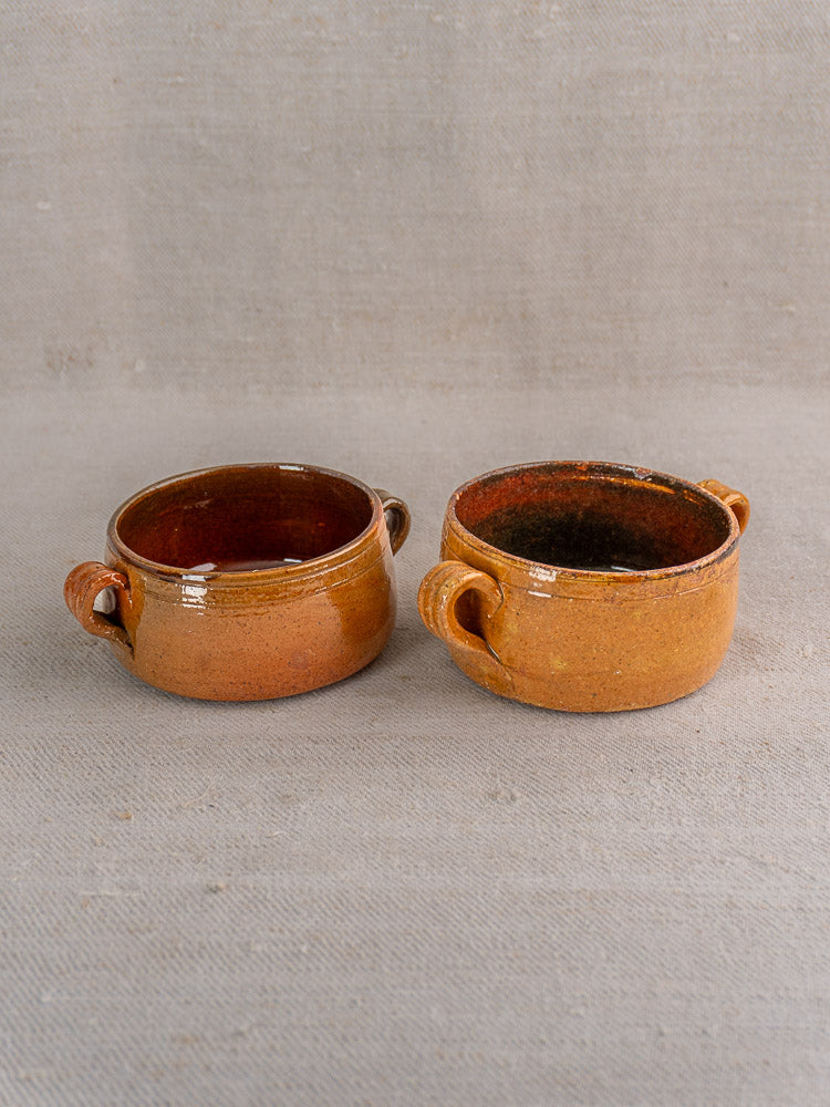 Pair of small round brown glazed terracotta French Vallauris bowls with side handles on beige surface.