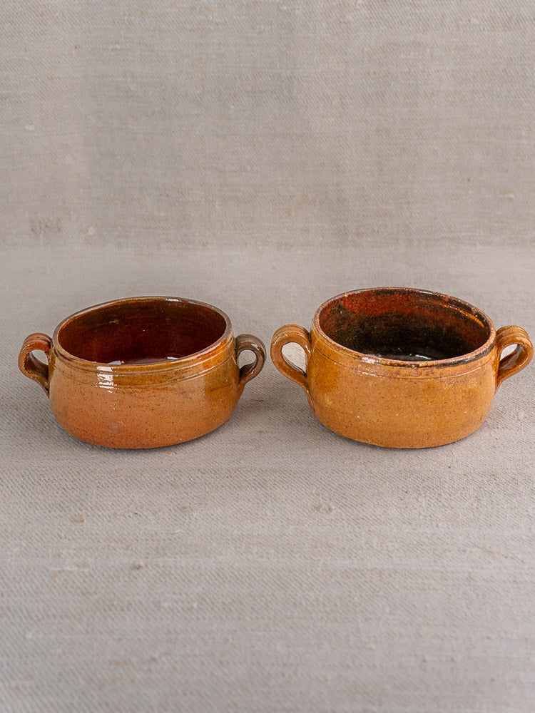 Pair of small, round brown glazed terracotta French Vallauris bowls with side handles on beige fabric.