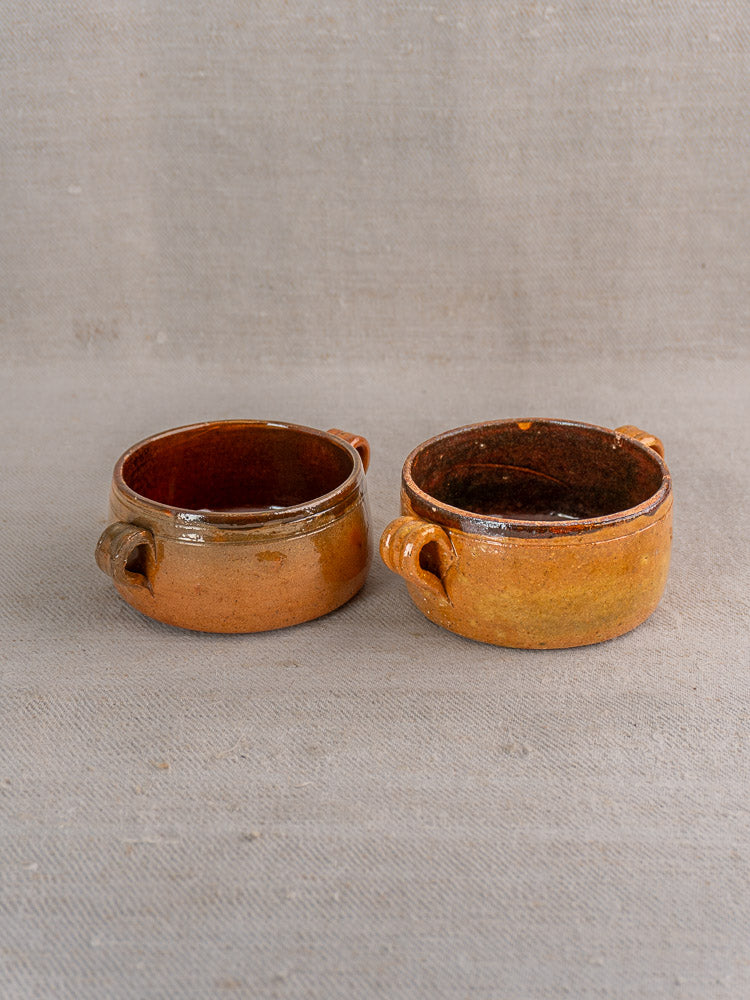 Set of three antique French Vallauris glazed terracotta pots with handles on gray fabric background.