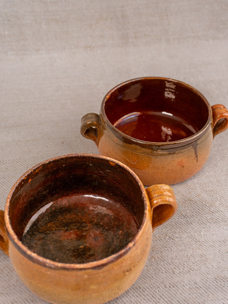 Set of three round, brown glazed French Vallauris terracotta pots with side handles and rustic finish on beige surface.