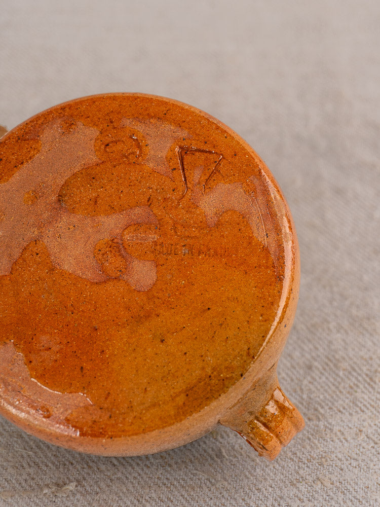 Round brown glazed ceramic Vallauris pot with two small handles, base markings, on textured fabric.