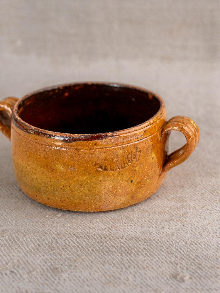 Small round brown glazed terracotta Vallauris bowl with two handles, darker interior, on beige textured surface.