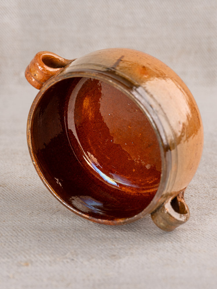 Brown glazed ceramic Vallauris vessel with two side handles, shown resting on textured light surface.