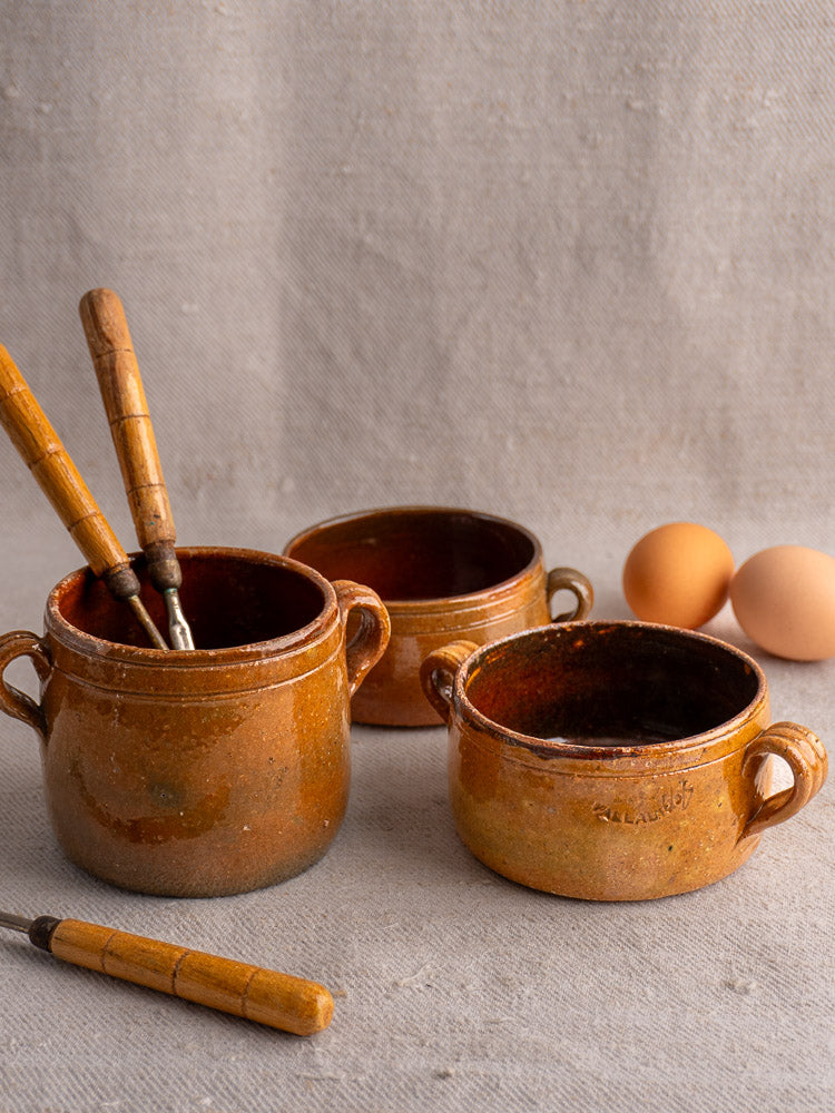 Three antique French Vallauris terracotta pots with wooden-handled utensils and two eggs on beige surface.
