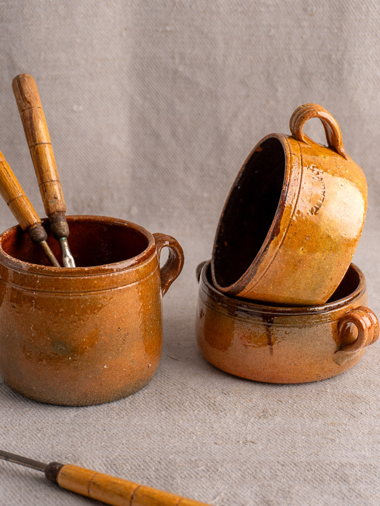 Set of three antique French Vallauris glazed terracotta pots with wooden-handled utensils on fabric surface.
