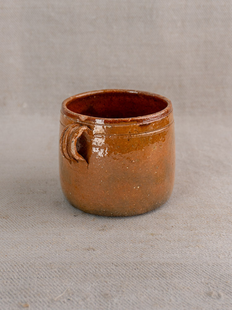 Handmade brown ceramic cup with glossy finish and small handle, inspired by antique French Vallauris pottery, on beige textured fabric.