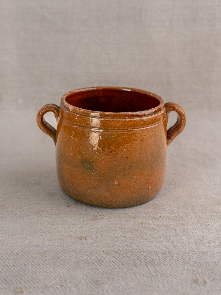 Small round brown glazed terracotta Vallauris pot with two handles on beige textured fabric.