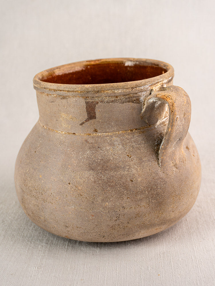 Antique French toupin cooking pot in rustic brown earthenware, two handles, glazed interior, on light fabric.