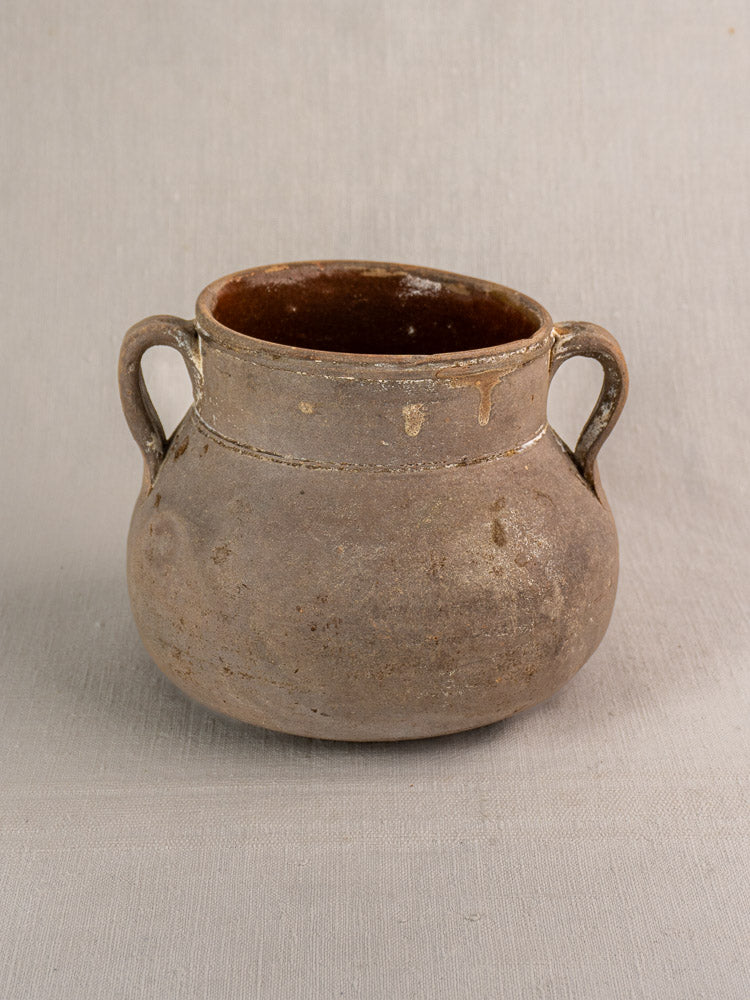 Antique French toupin, round brown earthenware pot with two handles on neutral fabric background, weathered country pottery charm.