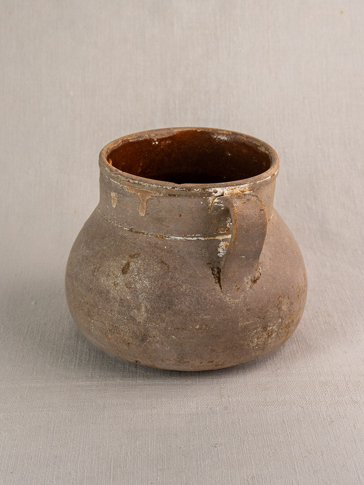Antique French earthenware toupin, aged brown with two handles and rustic patina, on light gray textured surface.