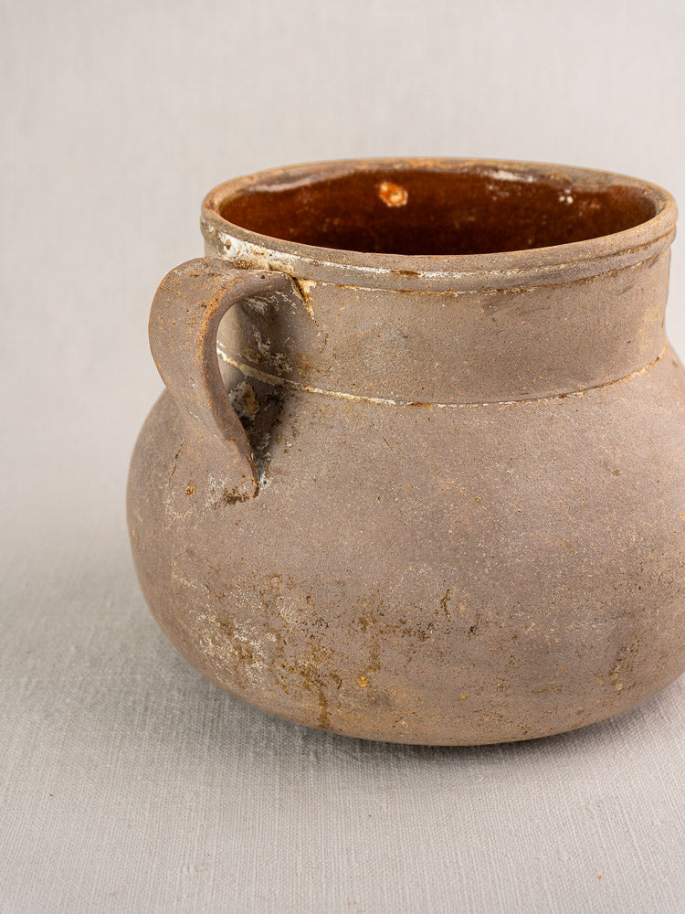 Antique French Toupin two-handle earthenware cooking pot with surface wear and light white residue on neutral fabric background.