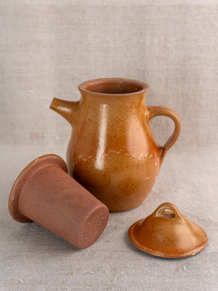 Antique French salt-glazed stoneware coffee pot with lid and infuser on textured beige fabric background.
