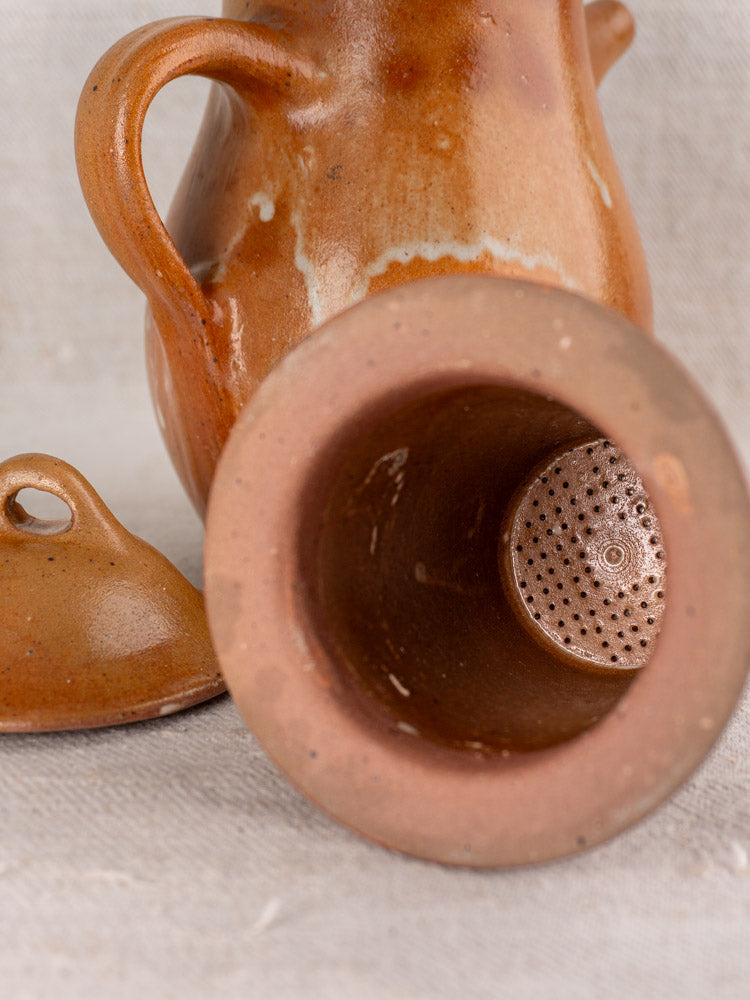 Antique French salt-glazed stoneware coffee pot with lid removed, showing strainer holes on a light textured surface.