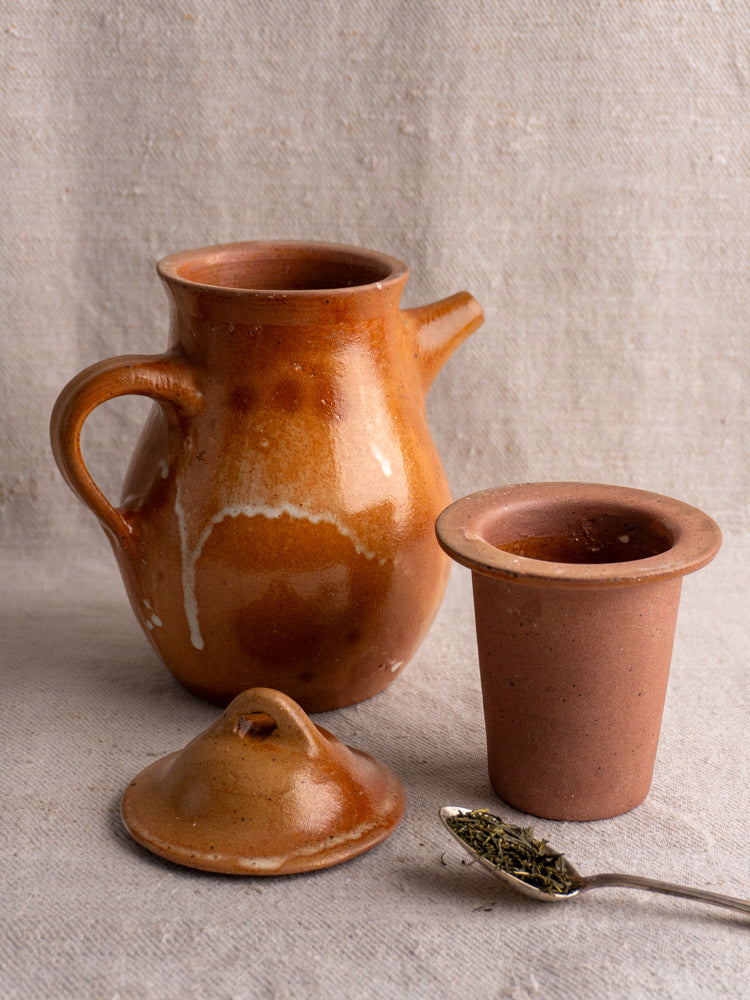 Antique French salt-glazed stoneware coffee pot with infuser, matching cup, lid, and spoon on neutral fabric.
