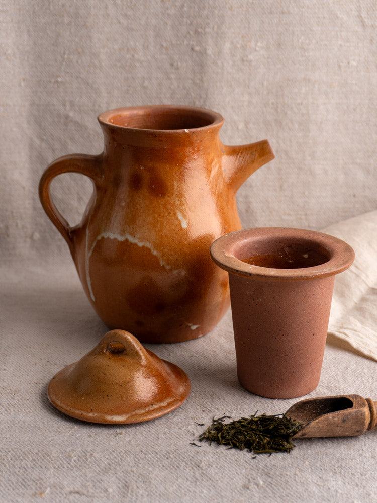 Antique French salt-glazed stoneware coffee pot with infuser, lid, cup, scoop, and tea leaves on neutral fabric. Mid 20th century.
