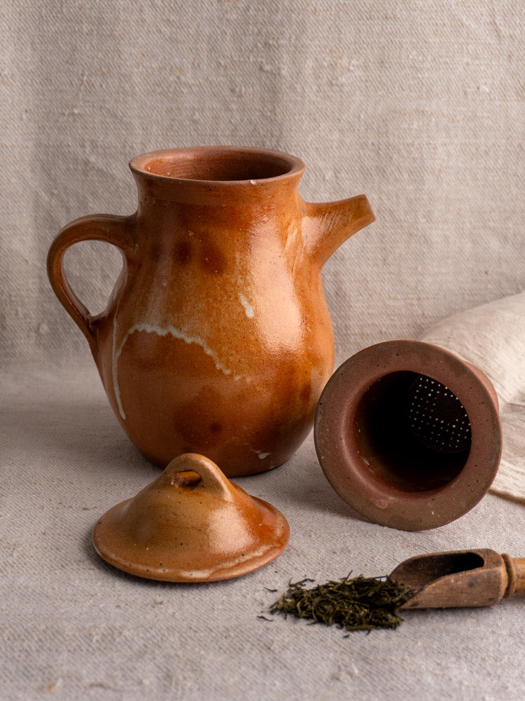 Antique French stoneware coffee pot in salt-glazed grès with infuser on beige fabric beside tea leaves and folded cloth.