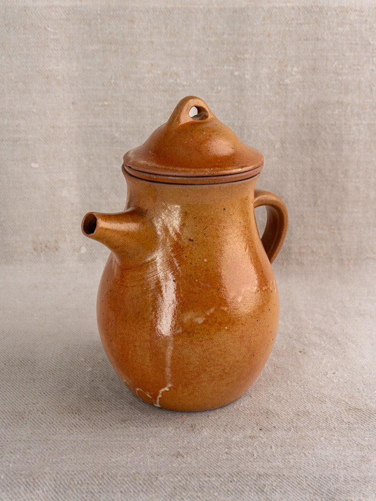 Antique French salt-glazed stoneware coffee pot with infuser and lid, glossy finish, small handle on neutral textured fabric.
