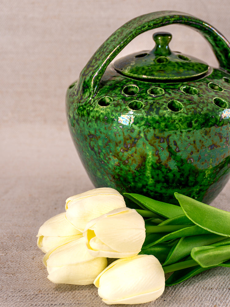 Antique French Vallauris green glazed chaufferette with lid holes beside white tulips on a textured neutral surface.