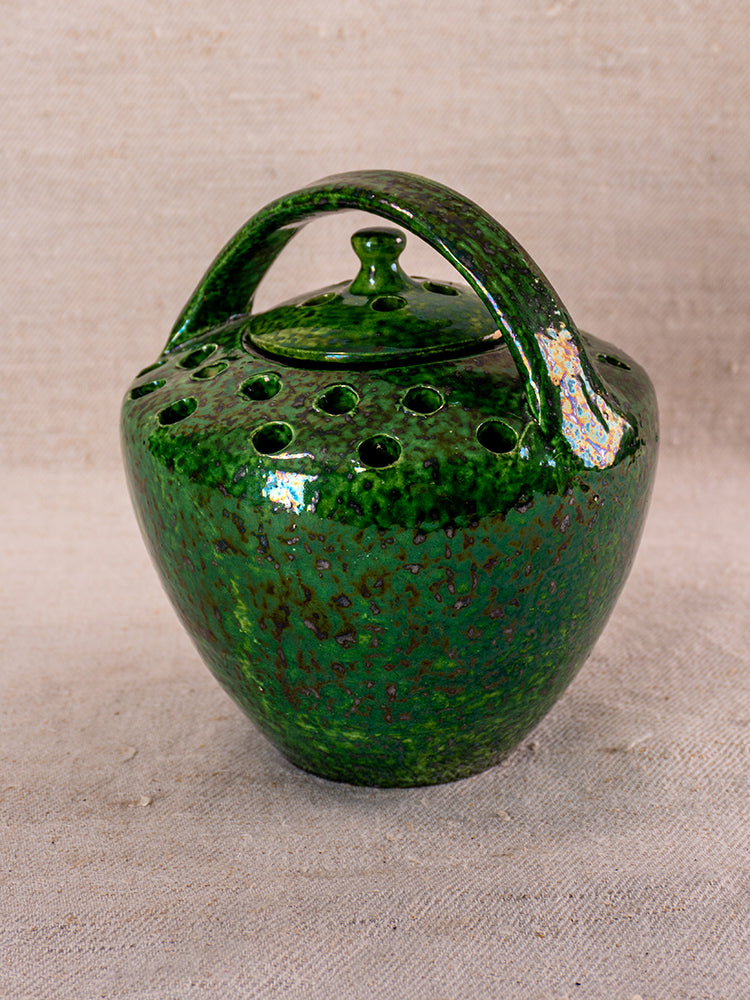 Antique French Vallauris green glazed chaufferette with rounded body, arched handle, lid, and perforated top on fabric background.