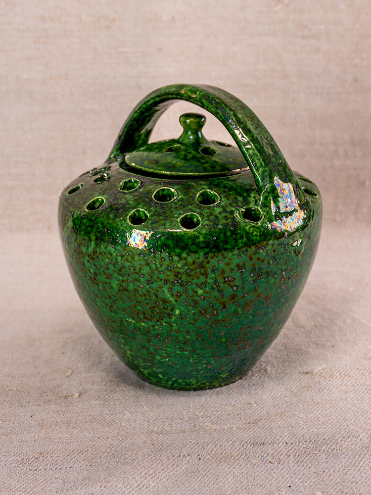 Antique French Vallauris green glazed ceramic chaufferette with lid and arched handle on textured fabric background.