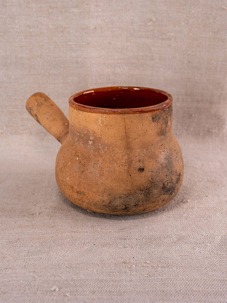 Antique Vallauris glazed terracotta pot à lait with single handle, reddish interior, on textured beige fabric.
