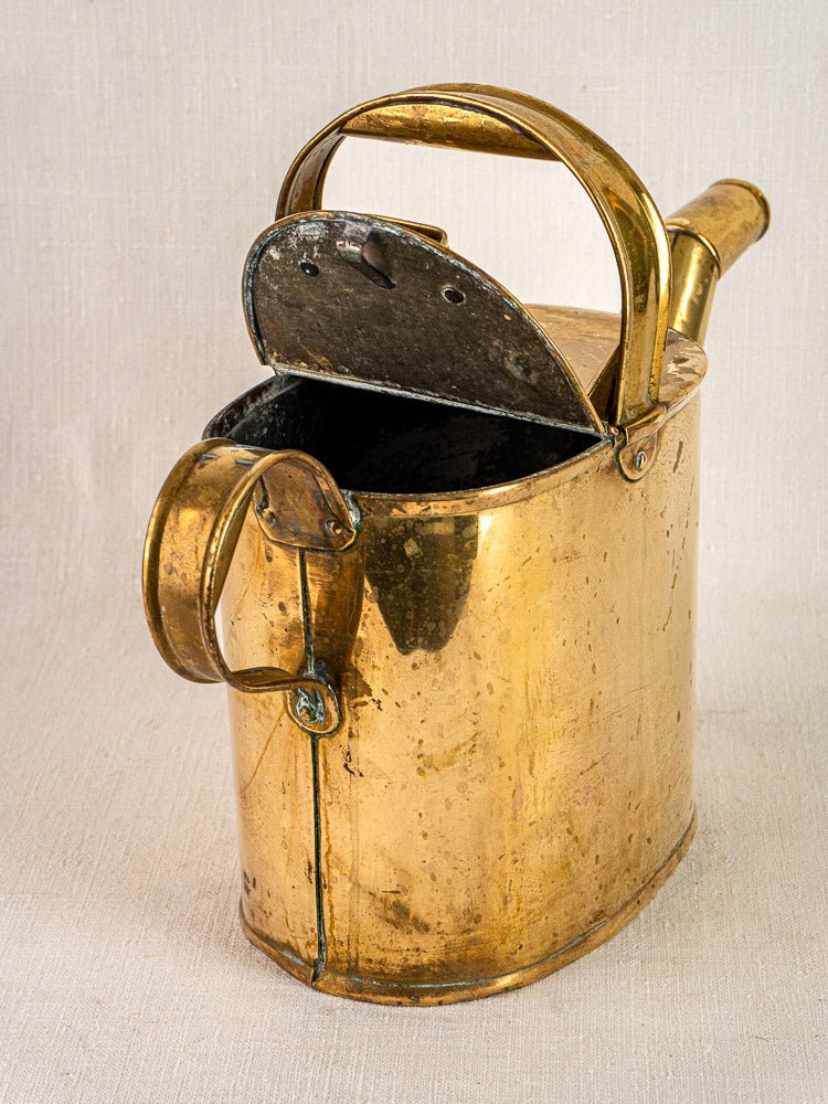 Antique French brass watering can with hinged lid and curved handle, displayed open on textured surface.