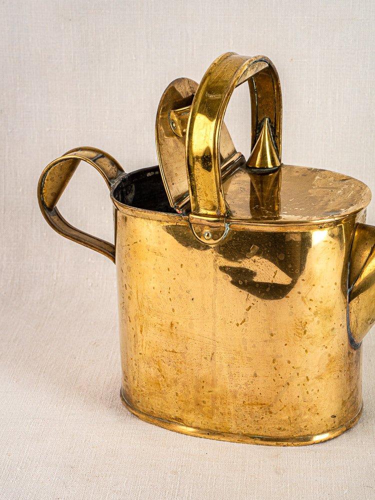 Antique French brass watering can with hinged lid, two handles, and spout on light fabric.