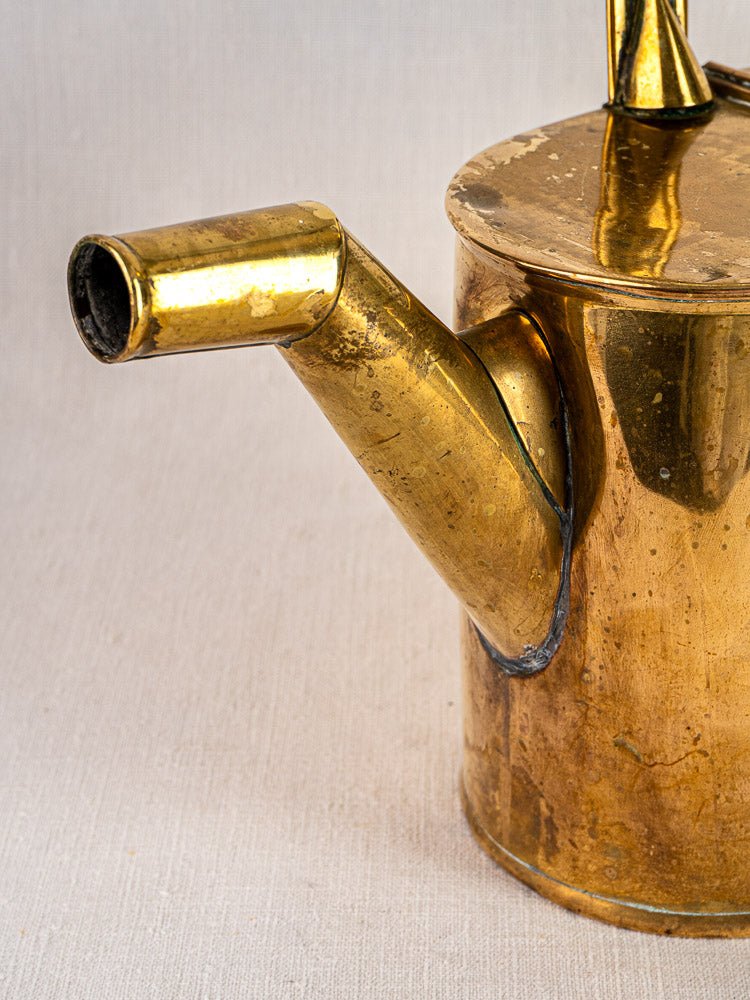 Antique French brass watering can with spout and hinged lid, shown on a neutral fabric background.