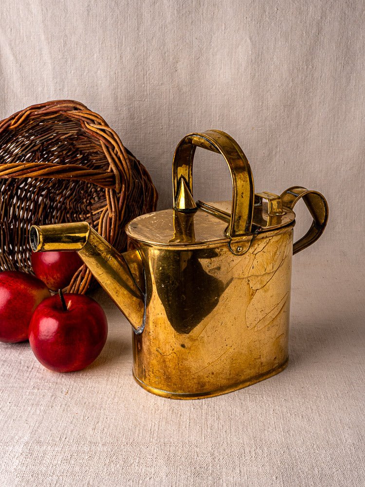 Antique French brass watering can with hinged lid, displayed on fabric, apples and basket behind.