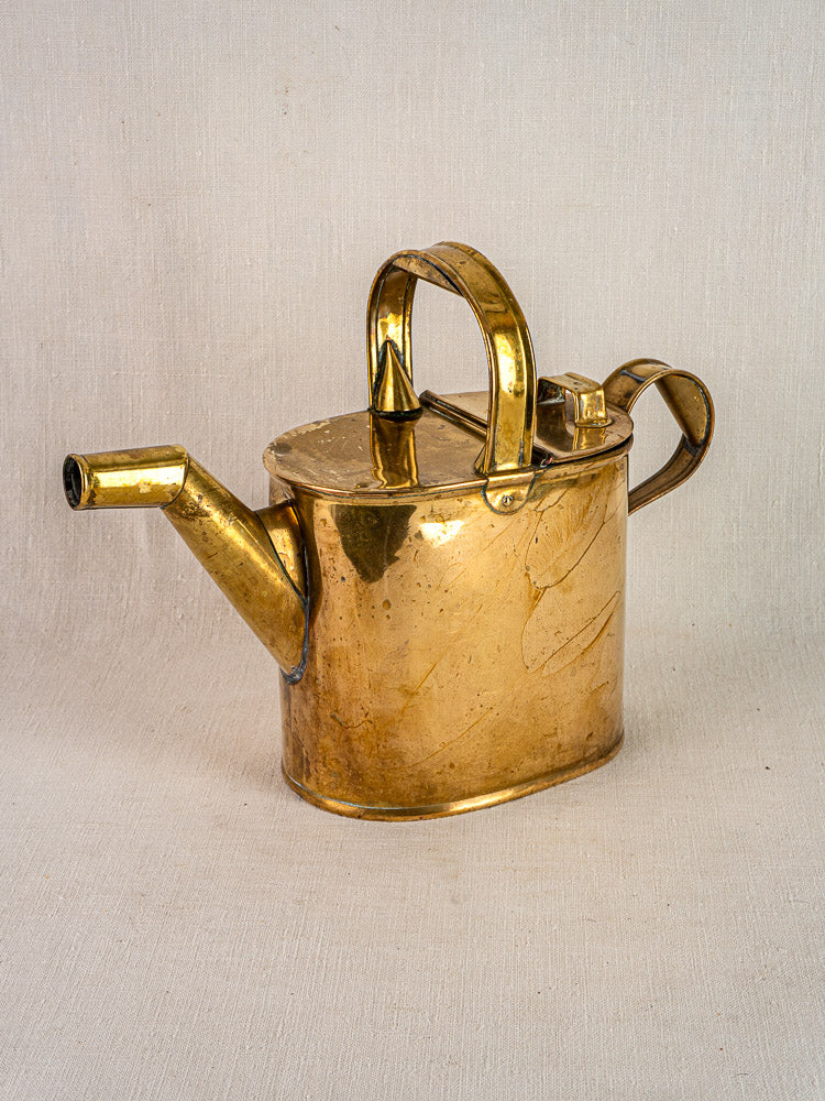 Antique French brass watering can with hinged lid and long spout on neutral fabric background.