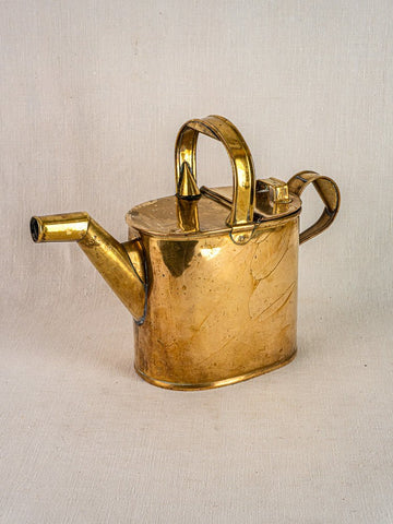 Antique French brass watering can with hinged lid and long spout on neutral fabric background.